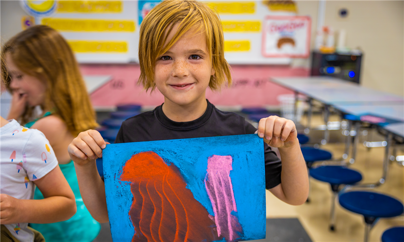 A boy with blond hair holds up a piece of art
